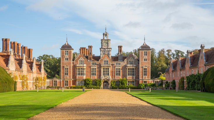 A red brick manor house stands at the end of a green lawn and is flanked by red brick buildings